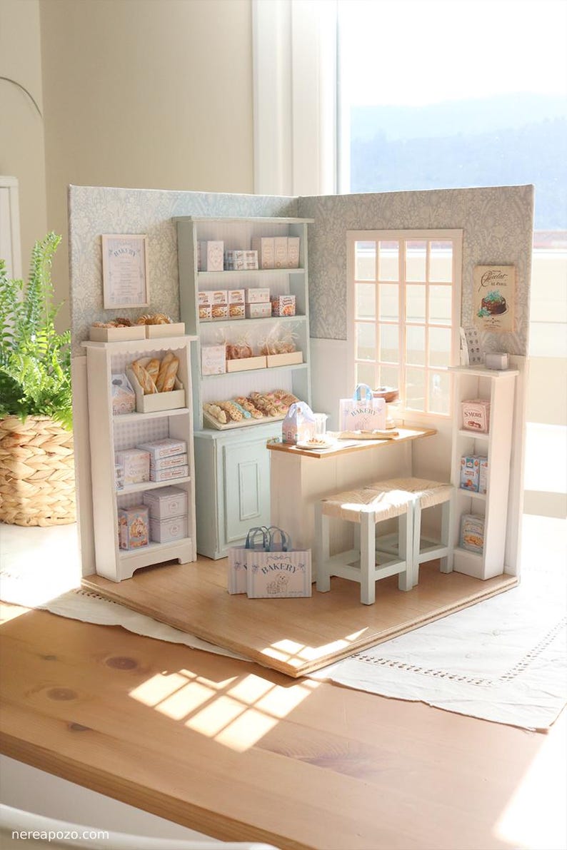 May include: A miniature bakery diorama featuring light blue and white wooden shelving units filled with tiny bread, pastries, and boxes. The scene includes a small counter with two stools, paper shopping bags labeled BAKERY, and wall art.