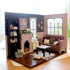 Könnte beinhalten: Eine Miniatur-Puppenstuben-Wohnzimmerszene mit dunklem Holzboden, einem schwarzen Bücherregal, einem hellvioletten Kamin mit Holzscheiten, einem weißen Beistelltisch, einem violett gemusterten Sofa mit verschiedenen Kissen und einem schwarzen Couchtisch.