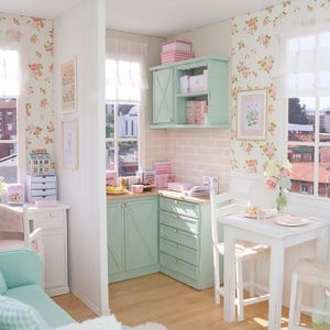 May include: A miniature kitchen with mint green cabinets and floral wallpaper. A small white table and chairs are set for a meal. The walls are decorated with framed artwork. The floor is light wood. The scene evokes a sense of a dollhouse.