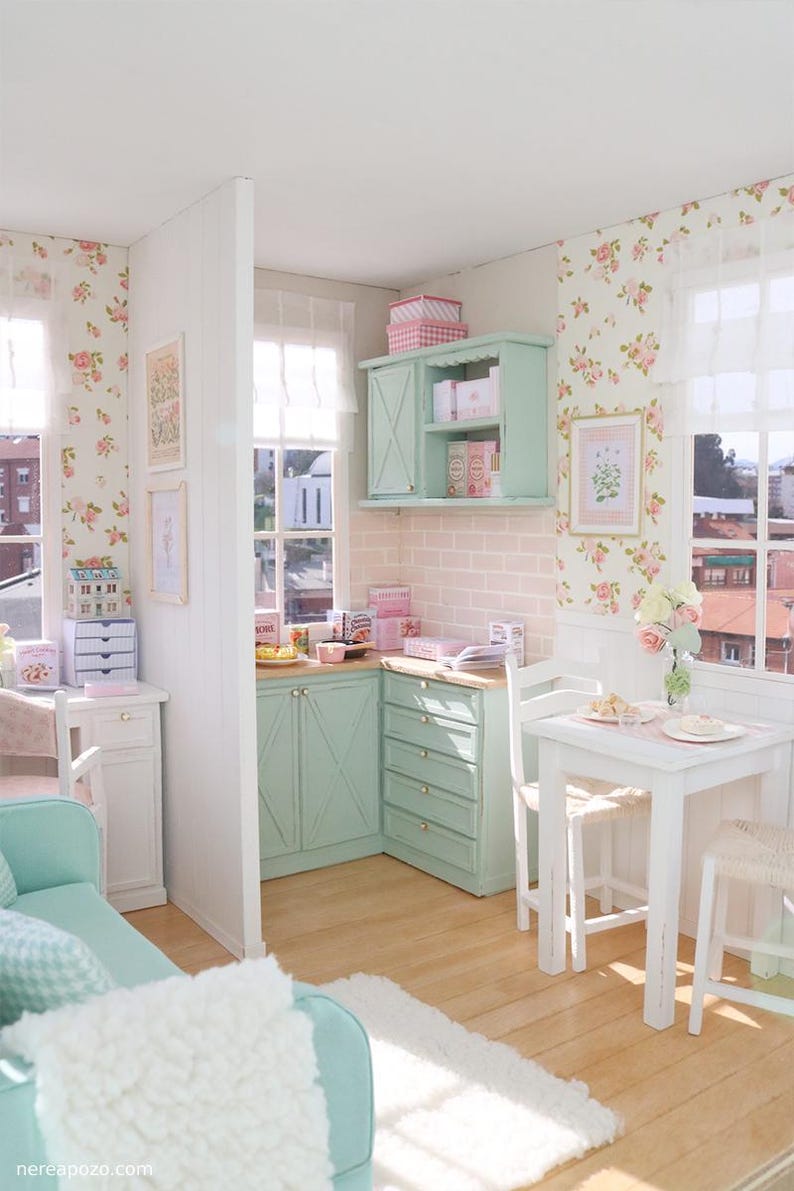 May include: A miniature kitchen with mint green cabinets and floral wallpaper. A small white table and chairs are set for a meal. The walls are decorated with framed artwork. The floor is light wood. The scene evokes a sense of a dollhouse.