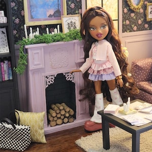 May include: A doll with long brown hair wearing a light pink cardigan, a layered pink and purple skirt, and white polka-dot leg warmers, standing in a miniature room with a light purple fireplace, a black table, and a checkered tote bag.