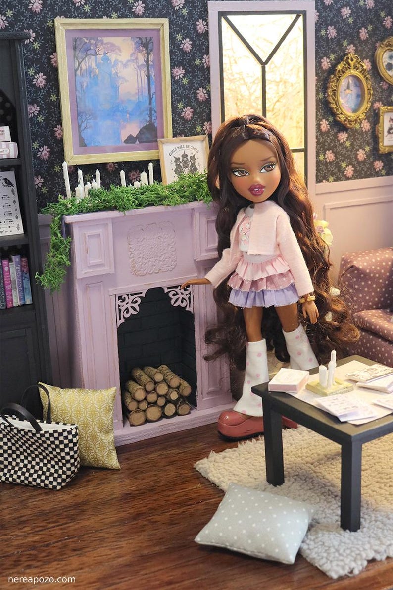 May include: A doll with long brown hair wearing a light pink cardigan, a layered pink and purple skirt, and white polka-dot leg warmers, standing in a miniature room with a light purple fireplace, a black table, and a checkered tote bag.