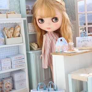 May include: A blonde doll with large eyes wearing a pink and white striped dress stands in a miniature bakery shop. The scene includes white shelves filled with boxes of cookies, bread, and two blue and white striped paper bags labeled BAKERY.