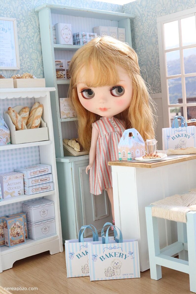 May include: A blonde doll with large eyes wearing a pink and white striped dress stands in a miniature bakery shop. The scene includes white shelves filled with boxes of cookies, bread, and two blue and white striped paper bags labeled BAKERY.