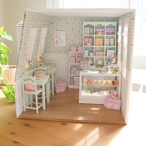K&ouml;nnte beinhalten: Miniatur-Diorama einer B&auml;ckerei und eines Caf&eacute;s in Hellgr&uuml;n und Rosa. Die Szene umfasst Tische, St&uuml;hle, Regale mit verpackten Waren und eine Vitrine mit Geb&auml;ck. Die W&auml;nde sind mit einem floralen Muster verziert.