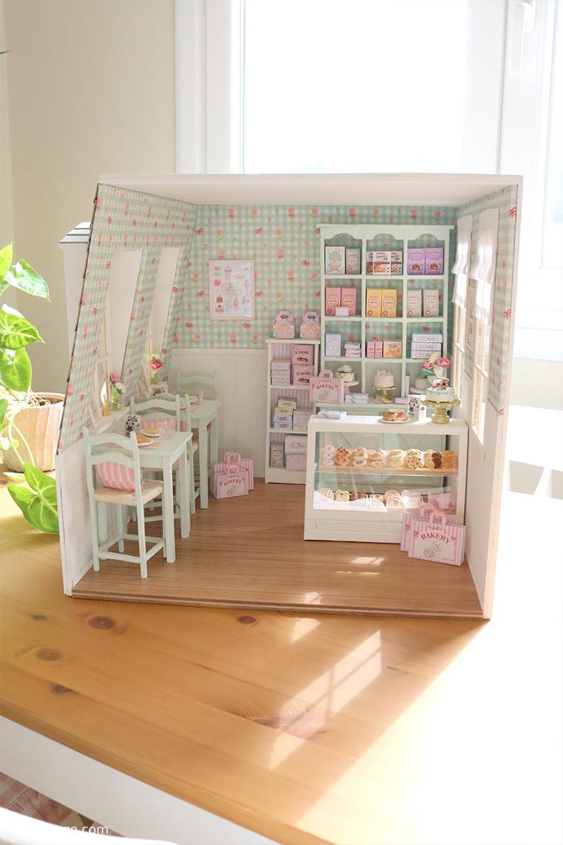 K&ouml;nnte beinhalten: Miniatur-Diorama einer B&auml;ckerei und eines Caf&eacute;s in Hellgr&uuml;n und Rosa. Die Szene umfasst Tische, St&uuml;hle, Regale mit verpackten Waren und eine Vitrine mit Geb&auml;ck. Die W&auml;nde sind mit einem floralen Muster verziert.