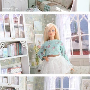 Może przedstawiać: Miniaturowe wnętrze domku dla lalek z białą szafką, książkami i oprawionymi dziełami sztuki. Lalka w bluzce w kwiaty i białej sp&oacute;dnicy znajduje się w pokoju. Pok&oacute;j ma okno z białymi zasłonami i ł&oacute;żko z poduszkami.