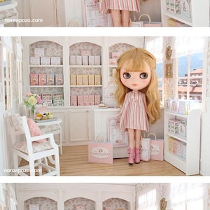 K&ouml;nnte beinhalten: Eine blonde Puppe mit gro&szlig;en Augen tr&auml;gt ein &auml;rmelloses Kleid mit vertikalen rosa, blauen und wei&szlig;en Streifen sowie rosa Stiefel und steht in einer Miniatur-B&auml;ckerei mit wei&szlig;en Regalen, pastellfarbenen Schachteln und Einkaufst&uuml;ten mit der Aufschrift Cake & Clover.