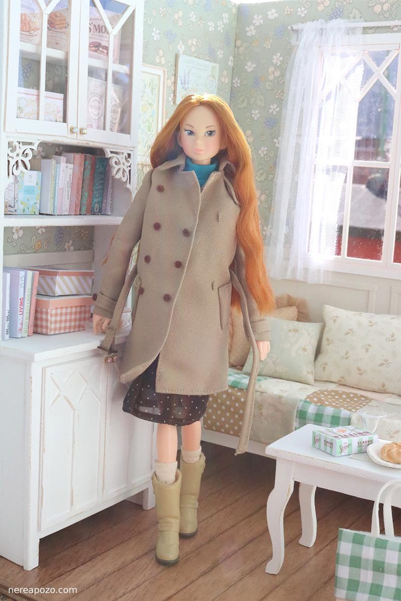 Może przedstawiać: Lalka z długimi, rudymi włosami w beżowym dwurzędowym płaszczu, ciemnej sp&oacute;dnicy w białe grochy i beżowych butach. Lalka stoi w miniaturowym pokoju z białą szafką, książkami i małym stolikiem.