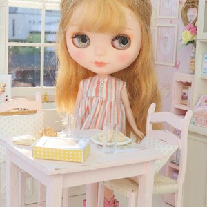 May include: A Blythe doll with blonde hair and rosy cheeks stands at a miniature pink table set for tea. The doll wears a striped dress and pink boots. The table has a yellow box with white spots, plates, and glasses.