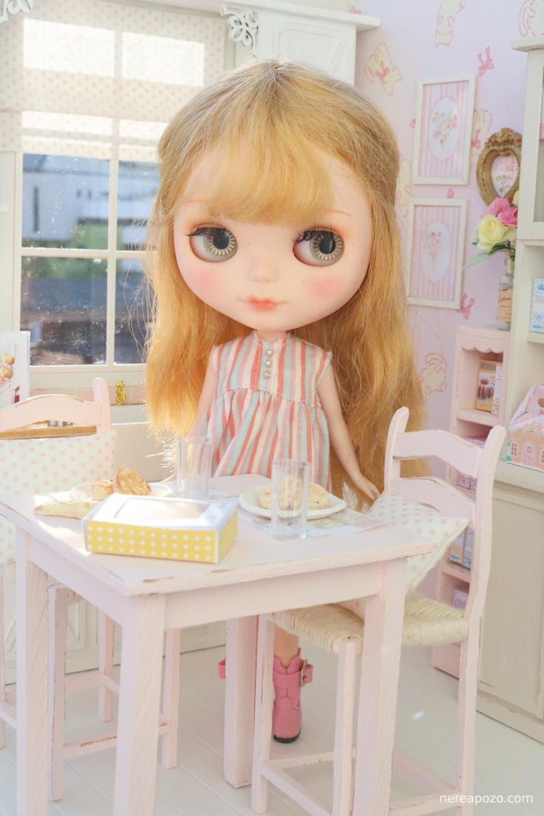 May include: A Blythe doll with blonde hair and rosy cheeks stands at a miniature pink table set for tea. The doll wears a striped dress and pink boots. The table has a yellow box with white spots, plates, and glasses.