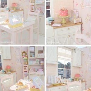 May include: A miniature doll's kitchen in pastel pink and white. The scene features a table, chairs, cabinets, and shelves stocked with miniature food and decorative items. A cake is on a gold stand, and the walls are decorated with framed pictures.