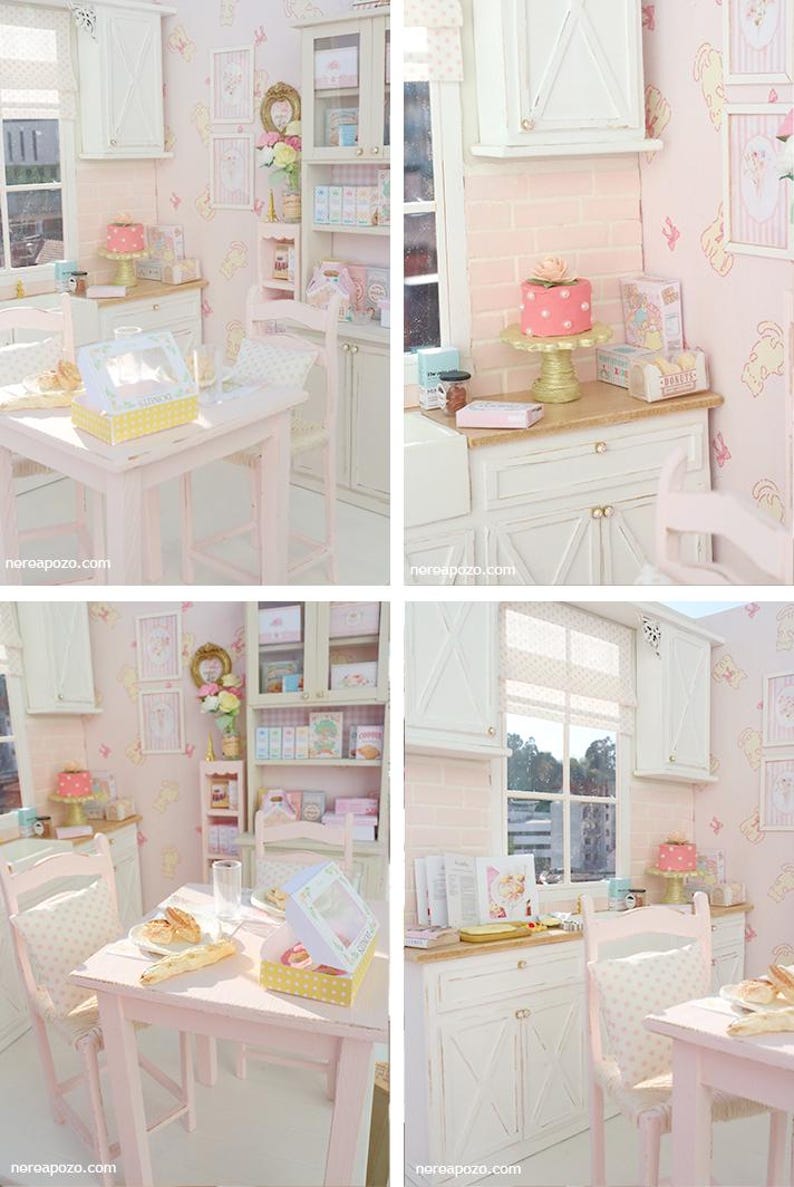 May include: A miniature doll's kitchen in pastel pink and white. The scene features a table, chairs, cabinets, and shelves stocked with miniature food and decorative items. A cake is on a gold stand, and the walls are decorated with framed pictures.