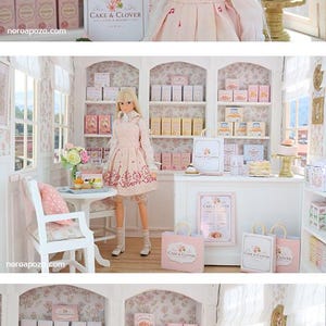 K&ouml;nnte beinhalten: Eine blonde Puppe in einem hellrosa Tr&auml;gerkleid und wei&szlig;er Bluse steht in einer Miniatur-B&auml;ckerei. Der Laden verf&uuml;gt &uuml;ber wei&szlig;e Regale mit kleinen Schachteln mit der Aufschrift Cake & Clover, eine wei&szlig;e Theke und Einkaufst&uuml;ten mit Logo.