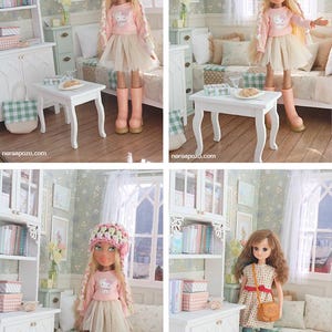 Może przedstawiać: Lalka w r&oacute;żowej bluzce z długim rękawem, beżowej sp&oacute;dnicy i r&oacute;żowych butach. Lalka ma r&oacute;żowo-zielony kapelusz z szydełka i długie blond warkocze. Lalka znajduje się w miniaturowym pokoju z białymi meblami i kwiatową tapetą.