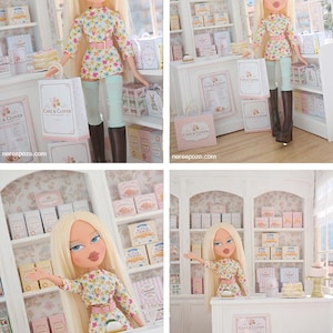K&ouml;nnte beinhalten: Eine blonde Puppe mit blauen Augen tr&auml;gt ein lang&auml;rmeliges Blumenoberteil, einen rosa G&uuml;rtel und hellblaue Leggings mit braunen Stiefeln. Die Puppe befindet sich in einer Miniatur-B&auml;ckerei mit wei&szlig;en Regalen, rosa-wei&szlig;en Schachteln und Cake & Clover-T&uuml;ten.