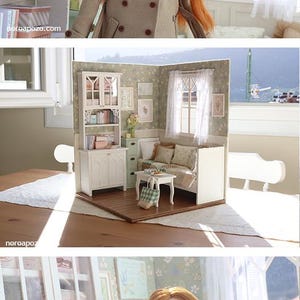 Może przedstawiać: Lalka z długimi rudymi włosami i beżowym płaszczem stoi w miniaturowym pokoju. Pok&oacute;j posiada białą biblioteczkę, małą sofę i okno z widokiem. Scena to szczeg&oacute;łowa miniatura diorama.