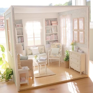 Könnte beinhalten: Miniatur-Puppenhaus-Wohnzimmer mit hellem Holzboden, einem beigen Sofa und einem weißen Sessel mit floralen Kissen. Der Raum verfügt über einen kleinen weißen Tisch, Bücherregale und eine weiße Kommode. Die Wände sind hellgrün gestrichen und mit floraler Tapete dekoriert.