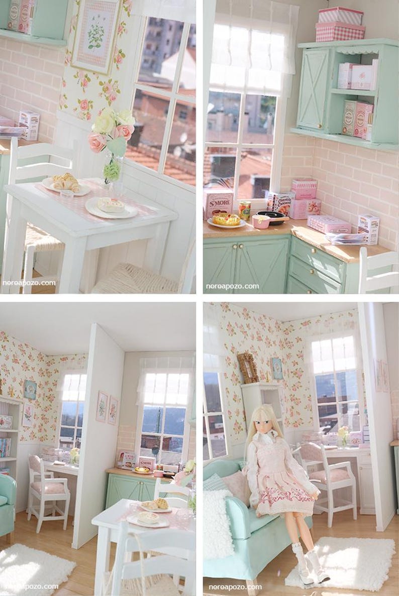 May include: A miniature dollhouse kitchen and dining room scene. The rooms feature white furniture, floral wallpaper, and pastel green cabinets. A doll is seated on a teal sofa. The scene includes a table setting, a window, and decorative items.