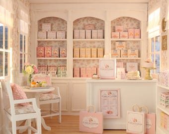 Könnte beinhalten: Eine Miniatur-Bäckerei-Auslage mit weißen Regalen voller verpackter Waren, einer Verkaufstheke mit Cake & Clover-Branding, Einkaufstaschen, einem kleinen Tisch mit zwei Stühlen und einer geblümten Tapete in einer sanften Pastellfarbpalette.