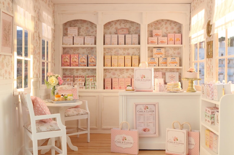 K&ouml;nnte beinhalten: Eine Miniatur-B&auml;ckerei-Auslage mit wei&szlig;en Regalen voller verpackter Waren, einer Verkaufstheke mit Cake & Clover-Branding, Einkaufstaschen, einem kleinen Tisch mit zwei St&uuml;hlen und einer gebl&uuml;mten Tapete in einer sanften Pastellfarbpalette.