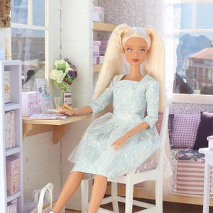 Pode incluir: Uma boneca com longos cabelos loiros em um vestido azul claro est&aacute; sentada em um quarto em miniatura. A boneca est&aacute; sentada em uma cadeira branca, com uma mesa e prateleiras por perto. Uma sacola e botas brancas tamb&eacute;m s&atilde;o vis&iacute;veis.