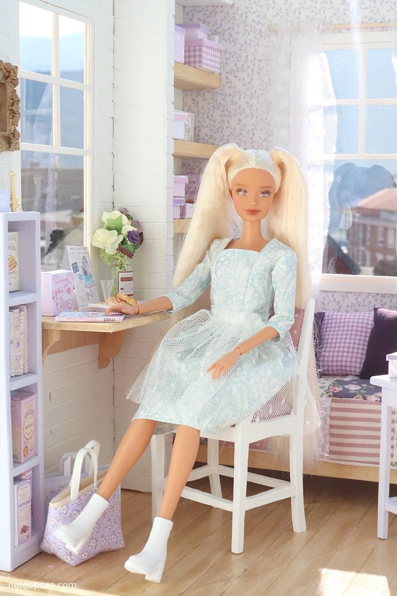 Pode incluir: Uma boneca com longos cabelos loiros em um vestido azul claro est&aacute; sentada em um quarto em miniatura. A boneca est&aacute; sentada em uma cadeira branca, com uma mesa e prateleiras por perto. Uma sacola e botas brancas tamb&eacute;m s&atilde;o vis&iacute;veis.