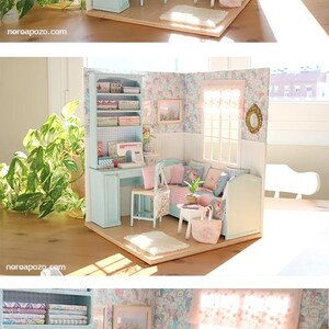 K&ouml;nnte beinhalten: Miniatur-Puppenhaus-Interieur mit N&auml;hbereich und Wohnzimmer. Der Raum ist in Pastellfarben gehalten und verf&uuml;gt &uuml;ber eine Blumtapete, eine N&auml;hmaschine und ein kleines Sofa mit Kissen. Das Gesamtthema ist eine gem&uuml;tliche, Vintage-&Auml;sthetik.