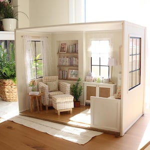 Könnte beinhalten: Ein Miniatur-Puppenhauszimmer mit einem beigefarbenen karierten Sessel und passendem Hocker, einem hölzernen Beistelltisch, eingebauten Bücherregalen mit winzigen Büchern, einem weißen Schrank mit Rattantüren, einer Stehlampe und weißen Vorhängen.