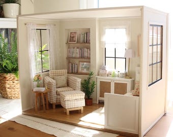 May include: A miniature dollhouse room featuring a beige checkered armchair and matching ottoman, a wooden side table, built-in bookshelves with tiny books, a white cabinet with cane doors, a floor lamp, and windows with white curtains.