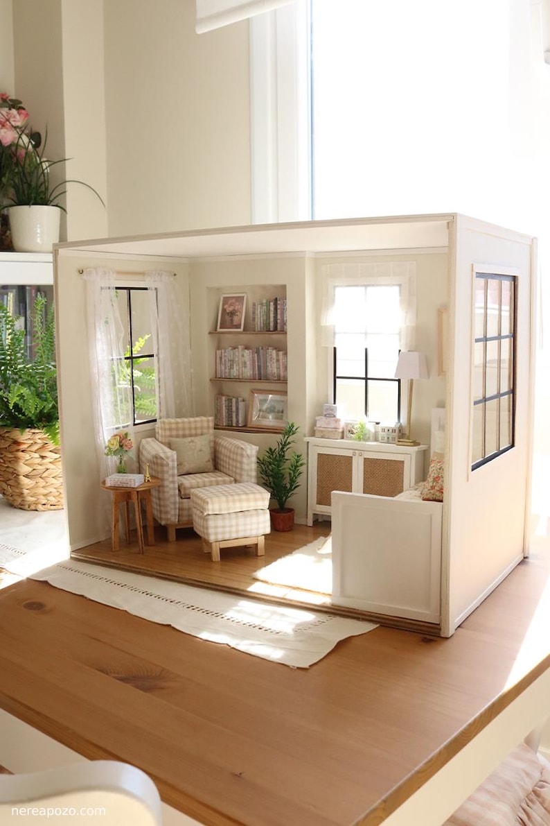 K&ouml;nnte beinhalten: Ein Miniatur-Puppenhauszimmer mit einem beigefarbenen karierten Sessel und passendem Hocker, einem h&ouml;lzernen Beistelltisch, eingebauten B&uuml;cherregalen mit winzigen B&uuml;chern, einem wei&szlig;en Schrank mit Rattant&uuml;ren, einer Stehlampe und wei&szlig;en Vorh&auml;ngen.