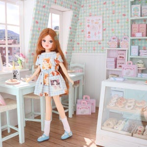 K&ouml;nnte beinhalten: Eine Puppe in einem hellblauen Kleid mit Katzenmotiven steht in einer Miniatur-B&auml;ckerei. Die B&auml;ckerei verf&uuml;gt &uuml;ber eine Vitrine mit Geb&auml;ck, Regale mit verpackten Waren und einen kleinen Tisch. Die W&auml;nde sind mit einem Blumenmuster verziert.