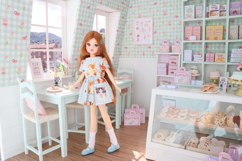 K&ouml;nnte beinhalten: Eine Puppe in einem hellblauen Kleid mit Katzenmotiven steht in einer Miniatur-B&auml;ckerei. Die B&auml;ckerei verf&uuml;gt &uuml;ber eine Vitrine mit Geb&auml;ck, Regale mit verpackten Waren und einen kleinen Tisch. Die W&auml;nde sind mit einem Blumenmuster verziert.