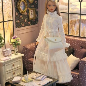 May include: A doll with long blonde hair wearing a long-sleeved white dress with a ruffled collar and tiered skirt. The doll carries a white crossbody bag with a gold-tone clasp. The doll is positioned in a room with a sofa and a small table.