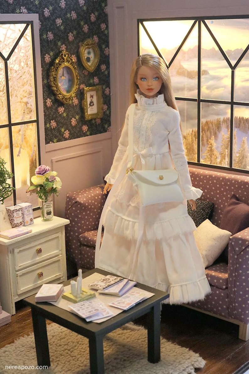 May include: A doll with long blonde hair wearing a long-sleeved white dress with a ruffled collar and tiered skirt. The doll carries a white crossbody bag with a gold-tone clasp. The doll is positioned in a room with a sofa and a small table.