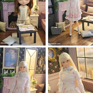 May include: A doll with long blonde hair wearing a white long-sleeved dress with a pink musical note and heart pattern, featuring a ruffled collar and lace trim. The doll stands in a miniature room setting with a fireplace and furniture.