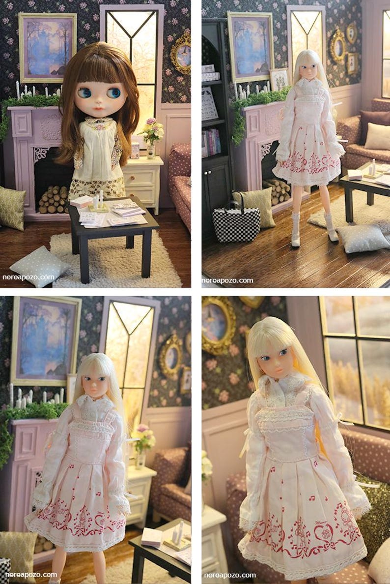 May include: A doll with long blonde hair wearing a white long-sleeved dress with a pink musical note and heart pattern, featuring a ruffled collar and lace trim. The doll stands in a miniature room setting with a fireplace and furniture.
