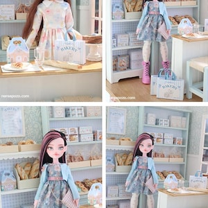 May include: A doll with long dark hair featuring pink highlights, wearing a floral dress, light blue cardigan, and pink boots. The doll stands in a miniature bakery setting with shelves filled with boxes, bread, and a sign labeled BAKERY.