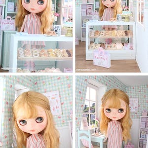 K&ouml;nnte beinhalten: Eine Puppe mit blonden Haaren und blauen Augen in einer Miniatur-B&auml;ckerei. Die Puppe tr&auml;gt ein gestreiftes Kleid und rosa Stiefel. Die B&auml;ckerei hat Regale mit Geb&auml;ck, eine Vitrine und einen Tisch mit einem Stuhl.