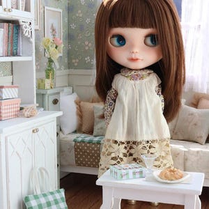 Może przedstawiać: Lalka Blythe z brązowymi włosami i niebieskimi oczami w miniaturowym pokoju. Ma na sobie kremową sukienkę z kwiatowym nadrukiem. Na białym stole znajduje się jedzenie i pudełko. Zielono-biała torba w kratę leży na podłodze.