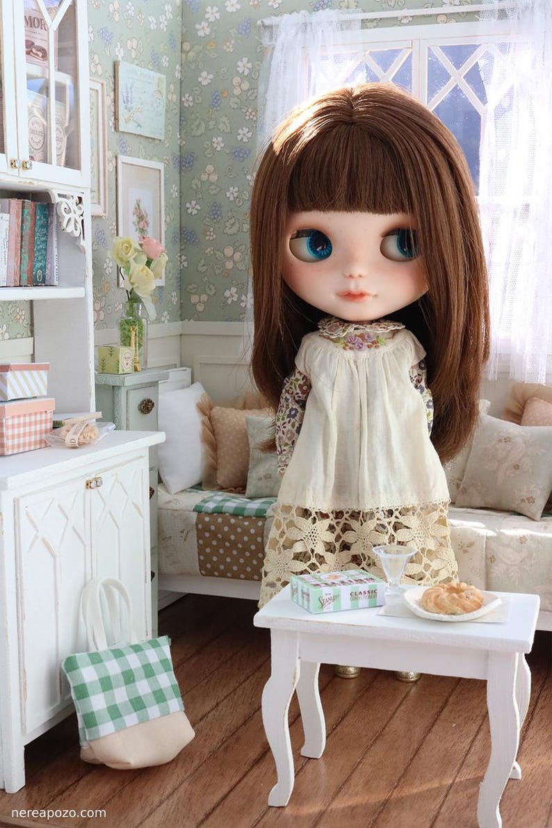 Może przedstawiać: Lalka Blythe z brązowymi włosami i niebieskimi oczami w miniaturowym pokoju. Ma na sobie kremową sukienkę z kwiatowym nadrukiem. Na białym stole znajduje się jedzenie i pudełko. Zielono-biała torba w kratę leży na podłodze.