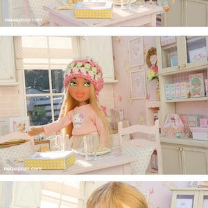 May include: A dollhouse kitchen scene featuring three dolls. One doll is in a blue cat-print dress, another in a pink sweater and hat, and the third in a striped dress. The kitchen has white cabinets, a window, and a table.