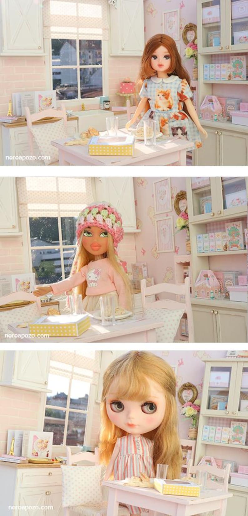 May include: A dollhouse kitchen scene featuring three dolls. One doll is in a blue cat-print dress, another in a pink sweater and hat, and the third in a striped dress. The kitchen has white cabinets, a window, and a table.