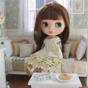 Może przedstawiać: Lalka z brązowymi włosami i niebieskimi oczami siedzi na miniaturowej sofie. Lalka ma na sobie kremową sukienkę z koronkowym wykończeniem. Mały biały stolik zawiera talerz z ciastkami i pudełko z napisem "Classic".