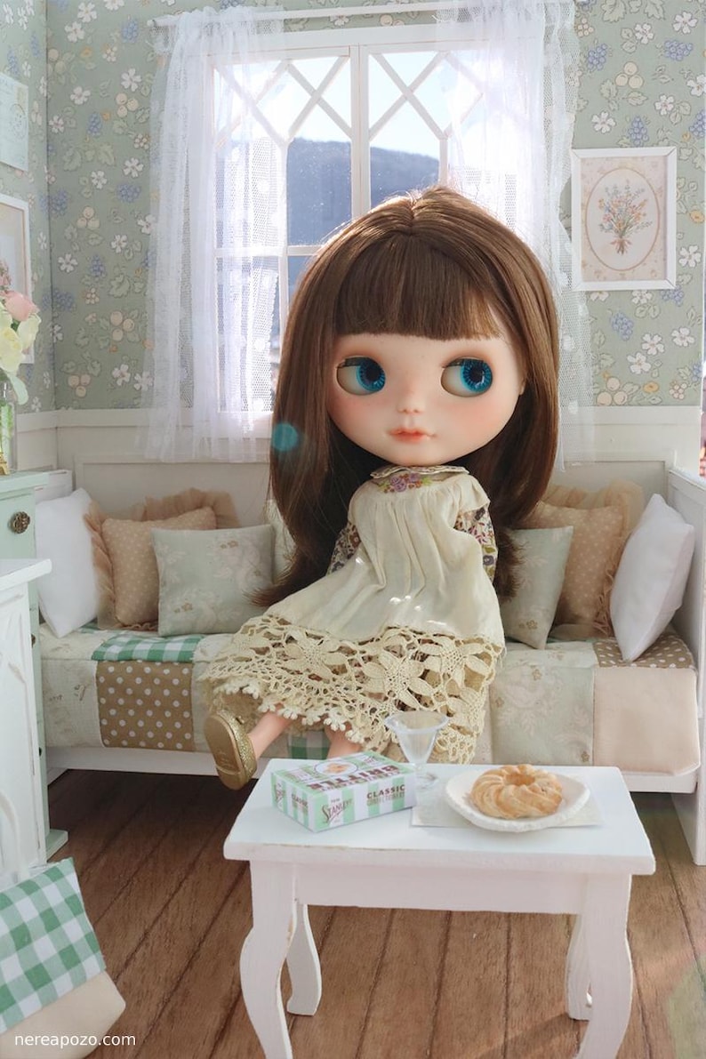 Może przedstawiać: Lalka z brązowymi włosami i niebieskimi oczami siedzi na miniaturowej sofie. Lalka ma na sobie kremową sukienkę z koronkowym wykończeniem. Mały biały stolik zawiera talerz z ciastkami i pudełko z napisem "Classic".