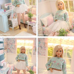 K&ouml;nnte beinhalten: Eine Puppe mit blonden Haaren, die ein hellblaues, gebl&uuml;mtes Crop-Top, einen wei&szlig;en T&uuml;llrock und rosa Leggings tr&auml;gt. Die Puppe befindet sich in einem Miniaturzimmer mit N&auml;hmaschine, Bett und Fenster. Der Raum ist in Pastellfarben gehalten.