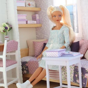 Pode incluir: Uma boneca loira com rabos de cavalo est&aacute; sentada em uma cena de quarto em miniatura. A boneca usa um vestido azul claro e botas brancas. O quarto apresenta uma cama, prateleiras com caixas e uma pequena mesa branca com uma caixa listrada.