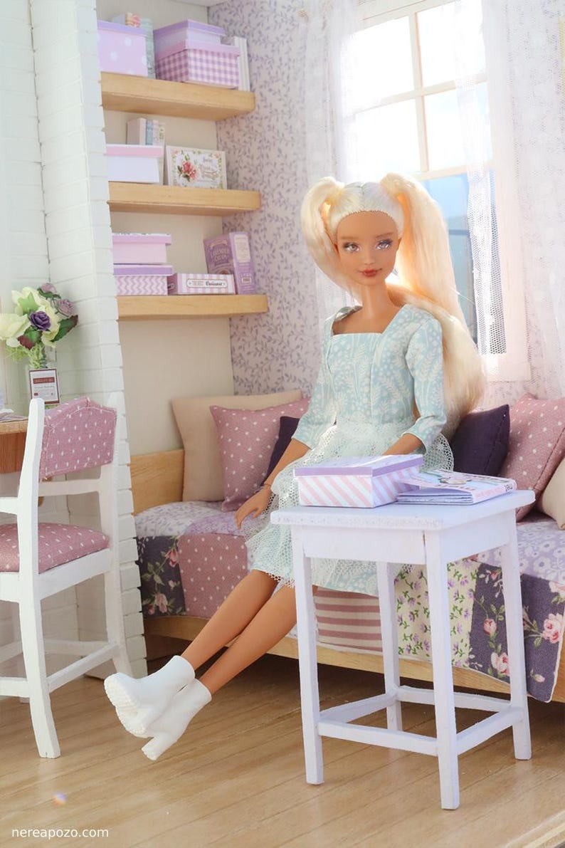 Pode incluir: Uma boneca loira com rabos de cavalo est&aacute; sentada em uma cena de quarto em miniatura. A boneca usa um vestido azul claro e botas brancas. O quarto apresenta uma cama, prateleiras com caixas e uma pequena mesa branca com uma caixa listrada.