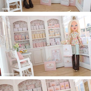K&ouml;nnte beinhalten: Eine Miniatur-Puppenhaus-B&auml;ckerei mit einer blonden Puppe mit langem Haar, die ein gebl&uuml;mtes Oberteil, eine hellblaue Leggings und dunkelbraune Stiefel tr&auml;gt. Der Laden enth&auml;lt wei&szlig;e Regale mit kleinen Schachteln, eine Theke und Einkaufst&uuml;ten mit der Aufschrift Cake & Clover.
