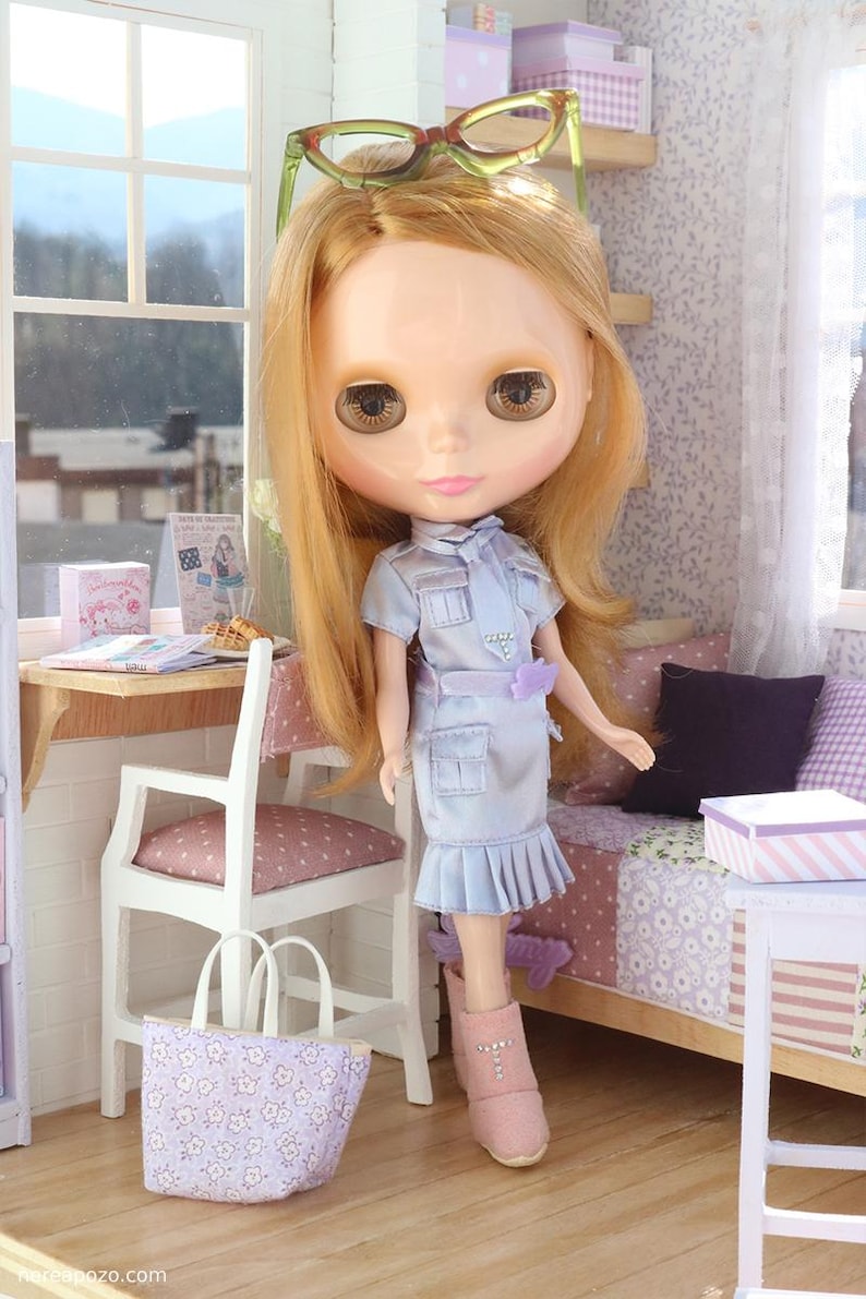 Pode incluir: Uma boneca Blythe com cabelo loiro comprido e olhos castanhos grandes, vestindo um vestido azul claro e botas rosa. A boneca tem &oacute;culos verdes na cabe&ccedil;a. Uma cadeira branca, uma sacola e um cen&aacute;rio em miniatura tamb&eacute;m s&atilde;o vis&iacute;veis.